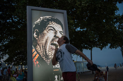 Tourists joke in front of an advertisement with the portrait of Uruguay's forward Luis Suarez at Copacabana beach in Rio de Janeiro, Brazil, on June 26, 2014. Sportswear giant Adidas said Thursday it would stop using Luis Suarez, one of its key promotional stars, for World Cup adverts after his four-month ban from football activities for biting Italian Giorgio Chiellini. AFP PHOTO / YASUYOSHI CHIBA 
